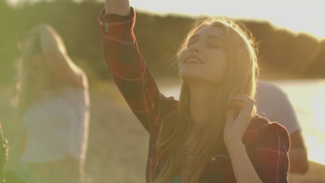 A-female-student-is-dancing-on-the-open-air-party-with-her-friends-on-the-beach.-Her-long-blonde-hair-is-flying-on-the-wind.-She-smiles-and-enjoys-a-summer-evening-on-the-river-coast-with-beer.